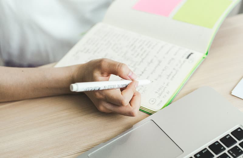 Dłoń zapisująca notatki w zeszycie obok laptopa